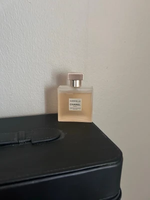 Chanel Gabrielle Eau de Parfum hår mist  - Elegant parfym från Chanel, Gabrielle Eau de Parfum hår mist, i en stilren fyrkantig glasflaska med ljusgul vätska och metallisk guldlock. Flaskan har en minimalistisk etikett med svart text. Perfekt för dig som gillar lyxiga dofter.