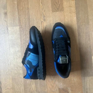 Valentino blå camo sneakers - Säljer ett par snygga sneakers från Valentino med blå och svarta camo-detaljer. Skorna har svarta snören, gummisula med nitar på hälen och är tillverkade i en mix av läder och textil. Perfekta för dig som vill sticka ut med en cool och unik look.