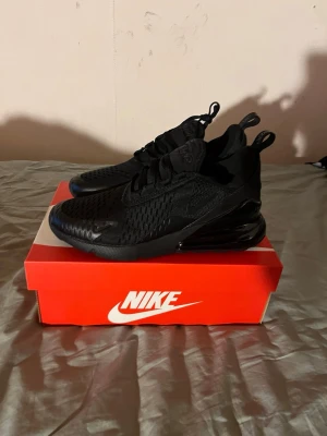 Nike Air Max 270 helsvarta sneakers - Nike Air Max 270 sneakers i helsvart med mesh-överdel och stora Air Max-dämpade sulor. Snygg sportig design med dragflikar på häl och plös, samt diskreta loggor. Perfekta för dig som gillar stilrena och bekväma sneakers.