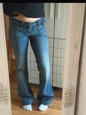 Blå bootcut jeans med låg midja - Säljer ett par blå bootcut jeans med låg midja och utsvängda ben. Jeansen har framfickor med knappdetaljer och klassisk femficksdesign. Perfekta för dig som gillar en retro och avslappnad stil. Materialet är denim i bomull.