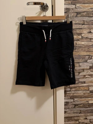 Svarta shorts från Tommy Hilfiger - Svarta shorts från Tommy Hilfiger med vit snörning i midjan och logotyp tryckt längs ena benet. Mjukt material och bekväm passform, perfekta för sommaren. Klassisk design med fickor både fram och bak.