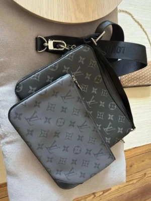  axelväska monogram svart - Snygg axelväska i svart med klassiskt monogrammönster. Väskan har justerbar axelrem, silverfärgade detaljer och en avtagbar innerficka med dragkedja. Tillverkad i slitstarkt canvasmaterial.