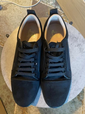 Svarta sneakers från Christian Louboutin - Stilrena svarta sneakers från Christian Louboutin med ovandel i mocka och klassiska svarta skosnören. Insidan är ljusbeige och märkt med guldfärgad logga. Snygg och minimalistisk design som passar till det mesta.