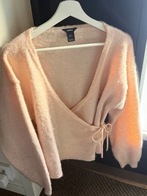 Ljusrosa omlottkofta från Lindex - Supermjuk ljusrosa omlottkofta från Lindex med knytband i midjan. Koftan har lång ärm, fluffig känsla och en avslappnad passform. Perfekt för lager-på-lager och ger en mysig vibe till din outfit.