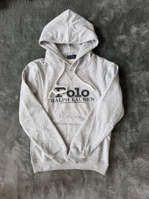 Grå hoodie från Polo Ralph Lauren - Snygg ljusgrå hoodie från Polo Ralph Lauren med stor logga framtill. Klassisk modell med huva och vita dragsnören. Tillverkad i mjukt bomullsmaterial och har ribbade muddar vid ärmslut och nederkant. Perfekt för en avslappnad och trendig stil.