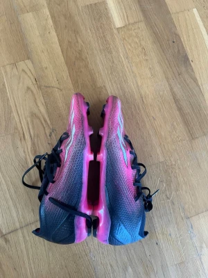 Svarta och rosa fotbollsskor Puma - Säljer ett par snygga fotbollsskor från Puma i svart och rosa med vita detaljer. Skorna har en modern, strömlinjeformad design och fasta dobbar för bra grepp på planen. Ovandelen är i syntetmaterial och snörningen ger bra passform. Perfekta för dig som vill sticka ut på planen.