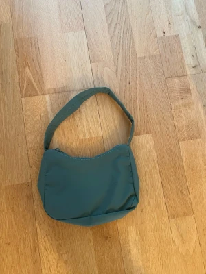 Grön axelväska - En stilren grön handväska med enkel design och mjuk form. Väskan har ett kort axelband och stängs med dragkedja upptill. Perfekt för dig som vill ha smidig väska till vardagen.