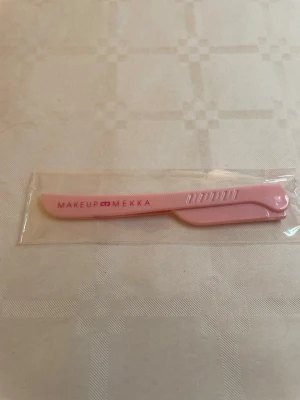 Rosa ögonbrynskniv från Makeup Mekka - En rosa ögonbrynskniv från Makeup Mekka i plast, perfekt för att forma och trimma ögonbryn eller ta bort små ansiktshår. Smidig och lätt att använda, med ett vikbart handtag för säker förvaring. Kommer i förpackning.