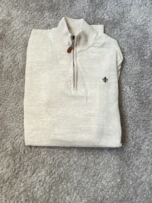 Beige halvzip-tröja från Morris - Stilren beige stickad tröja från Morris med halvzip och ribbad krage. Tröjan har en liten broderad logga på bröstet och dragkedja med läderdetalj. Perfekt för lager-på-lager och snygg till jeans eller chinos. En sjukt schysst tröja som jag använt enstaka gånger för att jag växt ur den tidigt.