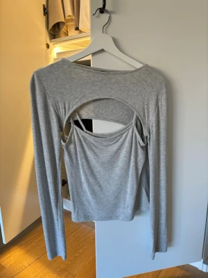 Grå långärmad topp med öppen rygg - Trendig grå långärmad topp med unik öppen rygg-detalj. Tillverkad i mjukt material som sitter skönt mot kroppen. Perfekt för dig som vill sticka ut med en cool och modern look.