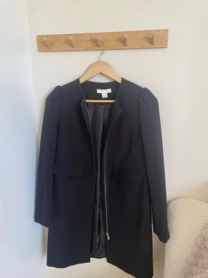 Svart kappa med dragkedja - Stilren svart kappa från HM med rak passform och diskreta axelvaddar. Kappan har en snygg guldfärgad dragkedja framtill och två fickor med lock. Perfekt för dig som gillar minimalistisk och klassisk stil.