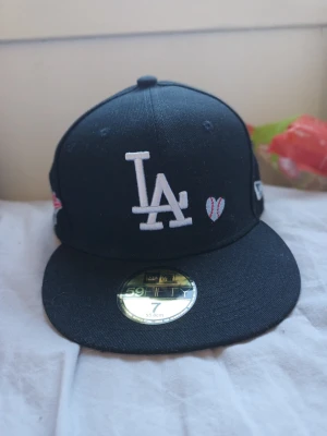 Svart LA Dodgers keps från New Era - Svart fitted keps 7 55.8 från New Era med LA Dodgers-logga broderad framtill och ett litet hjärta bredvid. Baksidan har texten 'Love + Hustle = Game' broderad i vitt och rött samt MLB-logga. Klassisk 59FIFTY-modell med platt skärm och cool streetkänsla.