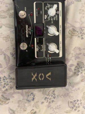 VOX StompLab SL1B multieffektpedal - Multieffektpedal från VOX, modell StompLab SL1B, i svart plast och metall med vita rattar och digital display. Pedalen har flera inställningar för olika musikstilar och effekter, samt en stor fotpedal med VOX-logotypen.