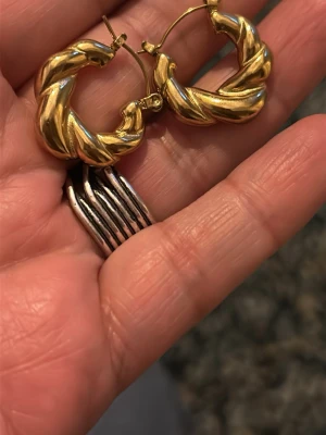 Guldiga vridna creolörhängen - Säljer ett par guldiga örhängen i creolmodell med vriden design. De är runda och chunky, vilket ger en trendig och iögonfallande look. Materialet ser ut att vara metall med guldplätering. Perfekta för dig som gillar statement-accessoarer.