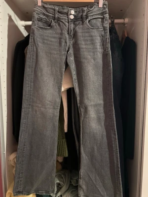 Grå bootcut jeans med dubbla knappar - Säljer ett par grå bootcut jeans med dubbla knappar framtill och klassiska fickor både fram och bak. Jeansen har en lätt tvättad look och är tillverkade i denim. Perfekt för dig som gillar en avslappnad och trendig stil.