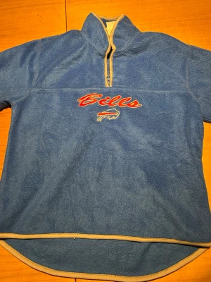 Blå Buffalo Bills fleece-tröja - Säljer en blå fleece-tröja med halv dragkedja och hög krage från Buffalo Bills. Tröjan har broderad logotyp och text på bröstet samt ljusa kantdetaljer. Perfekt för dig som gillar sport och vill ha något mjukt och bekvämt.