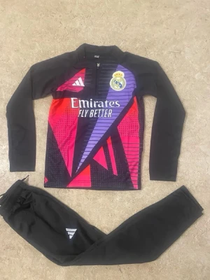 Real Madrid träningsset Adidas - Säljer ett Real Madrid träningsset från Adidas med långärmad tröja och byxor. Tröjan har svart bas med färgglada grafiska mönster i lila, rött och rosa samt klubbmärke och Adidas-logga. Byxorna är helsvarta med vit Adidas-detalj. Perfekt för fotbollsträning.