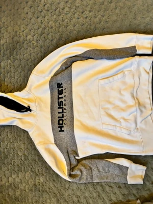 Vit och grå hoodie från Hollister - Snygg hoodie från Hollister i vitt och grått med svart logga på bröstet. Hoodien har huva med svarta snören och en stor magficka. Perfekt för en avslappnad och trendig stil. Mjuk insida och klassisk passform.
