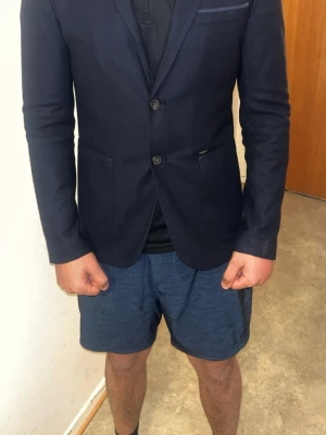 Mörkblå kavaj med armbågslappar - Snygg mörkblå kavaj med två knappar framtill och fickor på sidorna. Kavajen har coola mockadetaljer på armbågarna och en smal passform. Perfekt för dig som vill ha en stilren och modern look. Storlek S passar M