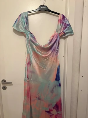 Pastellfärgad långklänning med blommor - Superfin långklänning i pastelliga färger med blommigt mönster. Klänningen har offshoulder-ärmar och en draperad urringning. Materialet är glansigt och känns som satin. Perfekt för sommarens alla tillfällen.