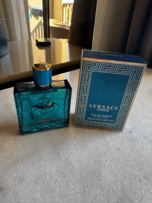 Versace Eros Eau de Parfum 100ml - Versace Eros Eau de Parfum i en snygg turkosblå glasflaska med guldiga detaljer och Medusa-logga. Flaskan är fyrkantig och kommer med originalkartong i matchande färger och mönster. En ikonisk doft från Versace som sticker ut i badrumshyllan.