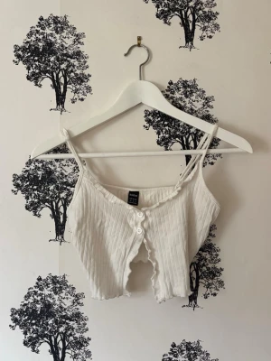 Vit ribbad croppad linnetopp - Säljer en vit ribbad croppad linnetopp från Shein med knappar framtill och smala axelband. Toppen har en snygg sliten kant och är perfekt för dig som gillar en trendig och avslappnad stil.