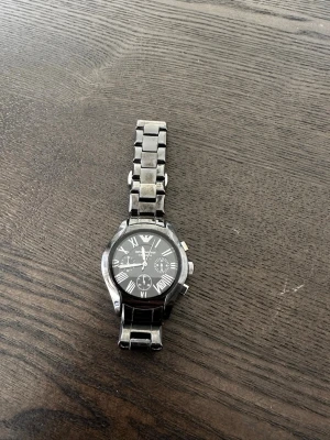 Emporio Armani svart klocka i metall - Stilren svart klocka från Emporio Armani med metallarmband och klassisk rund urtavla. Klockan har romerska siffror, flera små urtavlor och datumvisning. Perfekt accessoar för dig som gillar en elegant och tidlös look.