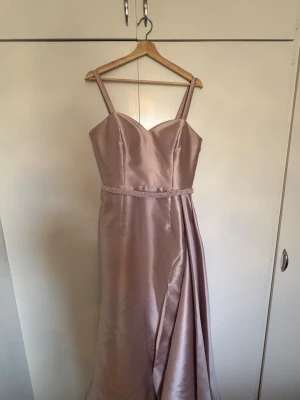 Ljusrosa långklänning med pärldetalj - Elegant ljusrosa långklänning i satin med smala axelband och vacker pärldetalj i midjan. Klänningen har en draperad effekt på ena sidan och en feminin v-ringning. Perfekt för speciella tillfällen och student. Tillkommer även ett vanligt skärp utan pärlor samt armpuffar. Klänningen har ett litet släp. 