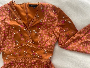 Orange blommig kortklänning från French Connection - Snygg orange kortklänning från French connection med blommigt mönster i rosa och vitt. Klänningen har v-ringning, spetsdetaljer och långa ärmar samt dragkedja i ryggen. Materialet känns som satin och ger ett lyxigt intryck. Perfekt för dig som vill sticka ut med färg och detaljer. Använd enstaka gånger, ordinarie pris ca 1500