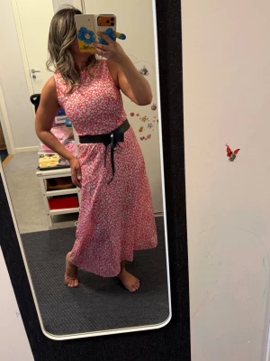 Rosa långklänning med svart bälte - Snygg rosa långklänning med vitt prickigt mönster och ett svart knytbälte i midjan. Klänningen är ärmlös och har en rund halsringning. Perfekt för dig som vill ha en cool och bekväm look. Materialet känns lätt och passar bra till varma dagar.helt ny