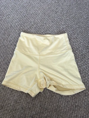 Ljusgula högmidjade shorts - Säljer ett par ljusgula högmidjade shorts i syntetmaterial. De har en clean och enkel design med bred resår i midjan och korta ben. Perfekta för träning eller chill dagar.
