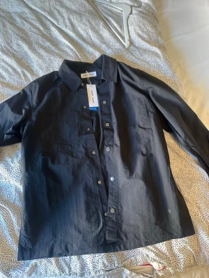 Svart overshirt från Arket - Stilren svart overshirt från samsoe  med klassisk krage, tryckknappar och två bröstfickor. Overshirten är tillverkad i ett lätt och strukturerat material som ger en modern look. Perfekt att bära öppen eller stängd över en t-shirt.