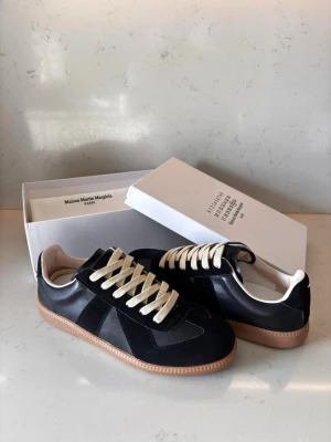 Maison Margiela  - Maison Margiela sneakers i svart läder och mocka med beigea snören och ljusbrun gummisula. Klassisk låg modell med stilren design och diskreta detaljer. Perfekt för dig som gillar minimalistiskt och trendigt mode. Skriv för frågor, pris kan diskuteras vid snabb affär.
