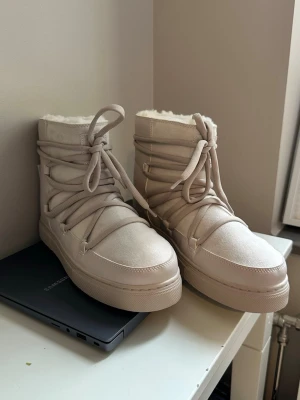 Beige moon boots med snörning - Säljer ett par beige moon boots från INUIKII med snörning och mjukt foder. Skorna har en rund tå, högt skaft och är tillverkade i ett läderliknande material med fluffigt innerfoder. Perfekta för kalla dagar och riktigt snygga till vinterlooken. Oanvänd och etiketten finns kvar 