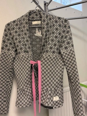 ODD MOLLY - Unik svartvit kofta från ODD MOLLY med traditionellt norskt mönster och långärmad design. Koftan har en rosa knytband framtill och är tillverkad i mjukt material, perfekt för dig som gillar stilrena och klassiska plagg med en twist. Storlek 1 passar S och M