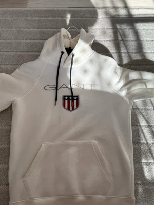 Vit hoodie från GANT med logga - Snygg vit hoodie från GANT med broderad logga och sköld på bröstet. Hoodien har huva med mörkblå dragsnören och en stor magficka framtill. Perfekt för en chill och stilren look.