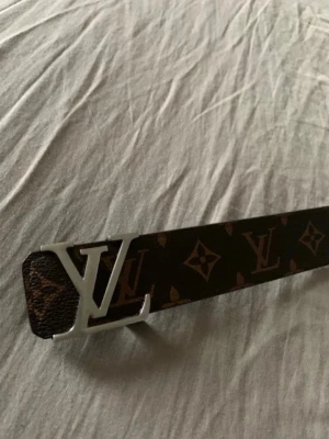  monogram bälte - Snyggt bälte med klassiskt monogrammönster i brunt och mörkbrunt. Bältet har ett spänne i silverfärgad metall och är tillverkat i skinn. Perfekt accessoar för att lyfta din outfit.