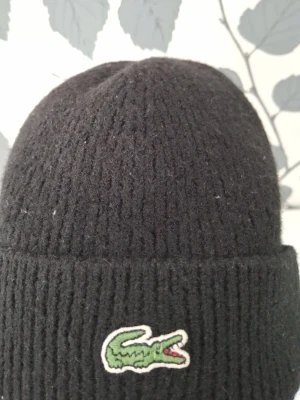 Svart stickad mössa från Lacoste - Svart ribbstickad mössa från Lacoste med uppvikt kant och den klassiska gröna krokodilloggan framtill. Tillverkad i mjukt och värmande material som håller dig varm under kalla dagar. Enkel och stilren design som funkar till det mesta.