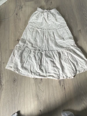 Vit långkjol i bomull med volanger - Säljer en luftig och somrig vit långkjol med flera volanglager. Kjolen har resår i midjan och är tillverkad i ett lätt bomullstyg som ger en avslappnad och flowy känsla. Perfekt för varma dagar och enkel att styla med olika toppar.