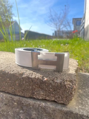 Vitt bälte med stor metallspänne - Snyggt vitt bälte med ett stort, blankt metallspänne i form av ett H. Bältet har en stilren och modern look som passar till många outfits. Tillverkat i skinn för en exklusiv känsla och lång hållbarhet.