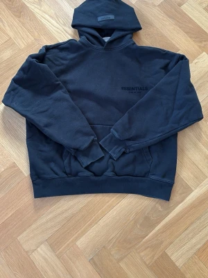 Svart Essentials Fear of God hoodie - Svart hoodie från Essentials Fear of God med diskret logga på bröstet och stor logga på ryggen. Klassisk känguruficka framtill, ribbade muddar och huva. Perfekt för en avslappnad streetwear-look. Kan köpa matchande byxor.