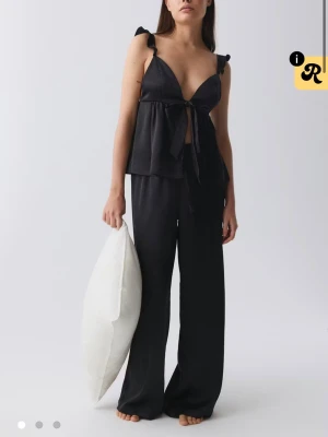 svart satinsätt med topp och byxa, pyjamas  - säljes ett svart satinsätt med en lös topp med knyt fram och breda axelband med volangdetaljer samt matchande vida byxor. Materialet är glansigt och mjukt, perfekt för en lyxig känsla. Snygg och avslappnad passform. använd fåtal gånger 