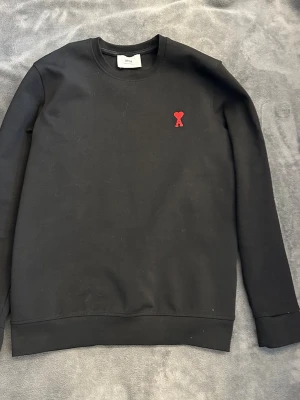 Svart stickad tröja från Ami Paris - Svart stickad tröja från Ami Paris med klassisk rund halsringning och lång ärm. På bröstet finns ett broderat rött hjärta med bokstaven A. Tröjan är enkel och stilren, perfekt för dig som gillar minimalistisk design och diskreta detaljer.