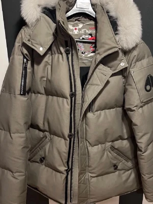 Moose Knuckles dunjacka med päls - Säljer en beige dunjacka från Moose Knuckles med avtagbar huva och lyxig ljus päls. Jackan har flera fickor med dragkedja, logotyp på ärmen och quiltad design. Perfekt för kalla vinterdagar och riktigt snygg att bära.