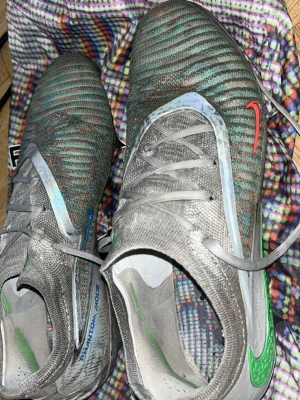 Nike Phantom fotbollsskor elite grå/grön - Nike Phantom Elite fotbollsskor med grå och gröna nyanser, detaljer i rött och blått samt grönt Swoosh-märke. Skorna har snörning och är tillverkade i syntetmaterial med textilstruktur för bra grepp och kontroll på planen. Perfekta för dig som vill ha stil och funktion på fotbollsplanen.