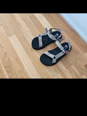 Beige sandaler från Tommy Hilfiger - Snygga sandaler från Tommy Hilfiger med beige justerbara remmar och svart sula. Klassisk design med logotyp på remmarna. Perfekta för sommaren och lätta att matcha till olika outfits.