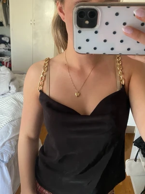 Svart linnetopp med guldkedjor - Snygg svart linnetopp med breda guldfärgade kedjor som axelband. Toppen har en lätt draperad urringning och är tillverkad i ett glansigt material som ger en lyxig känsla. storlek Xs, använd en gång 