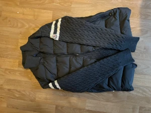 Svart pufferjacka från Moncler - Snygg svart pufferjacka från Moncler med stickade ärmar och två vita ränder på ena ärmen. Jackan har dragkedja och Moncler-logga på bröstet. Perfekt för kalla vinterdagar och riktigt trendig stil.