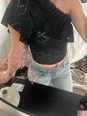 Svart one shoulder topp med volang - Säljer en svart one shoulder topp med volang och broderade hål. Toppen är croppad och har en asymmetrisk design med en axel bar. Perfekt för dig som vill sticka ut lite extra.