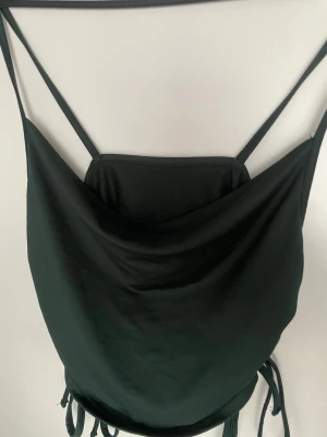 Mörk grön linnetopp med tunna axelband - Stilren mörk grön linnetopp med tunna axelband och draperad front. Toppen har knytband i sidorna och är perfekt för en trendig look. Materialet känns mjukt och följsamt mot huden.